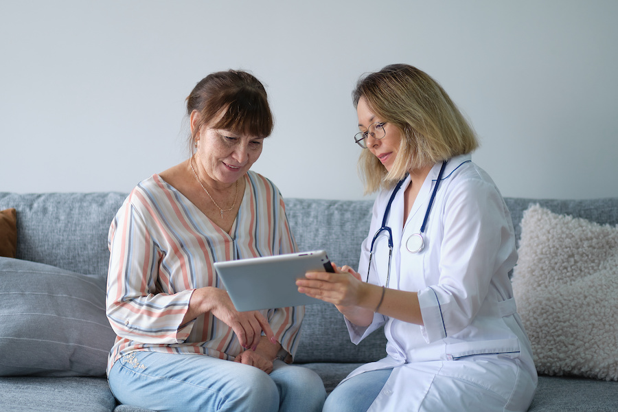 Female professional doctor showing medical test result explaining prescription using digital tablet app visiting senior woman patient at home sitting on sofa.